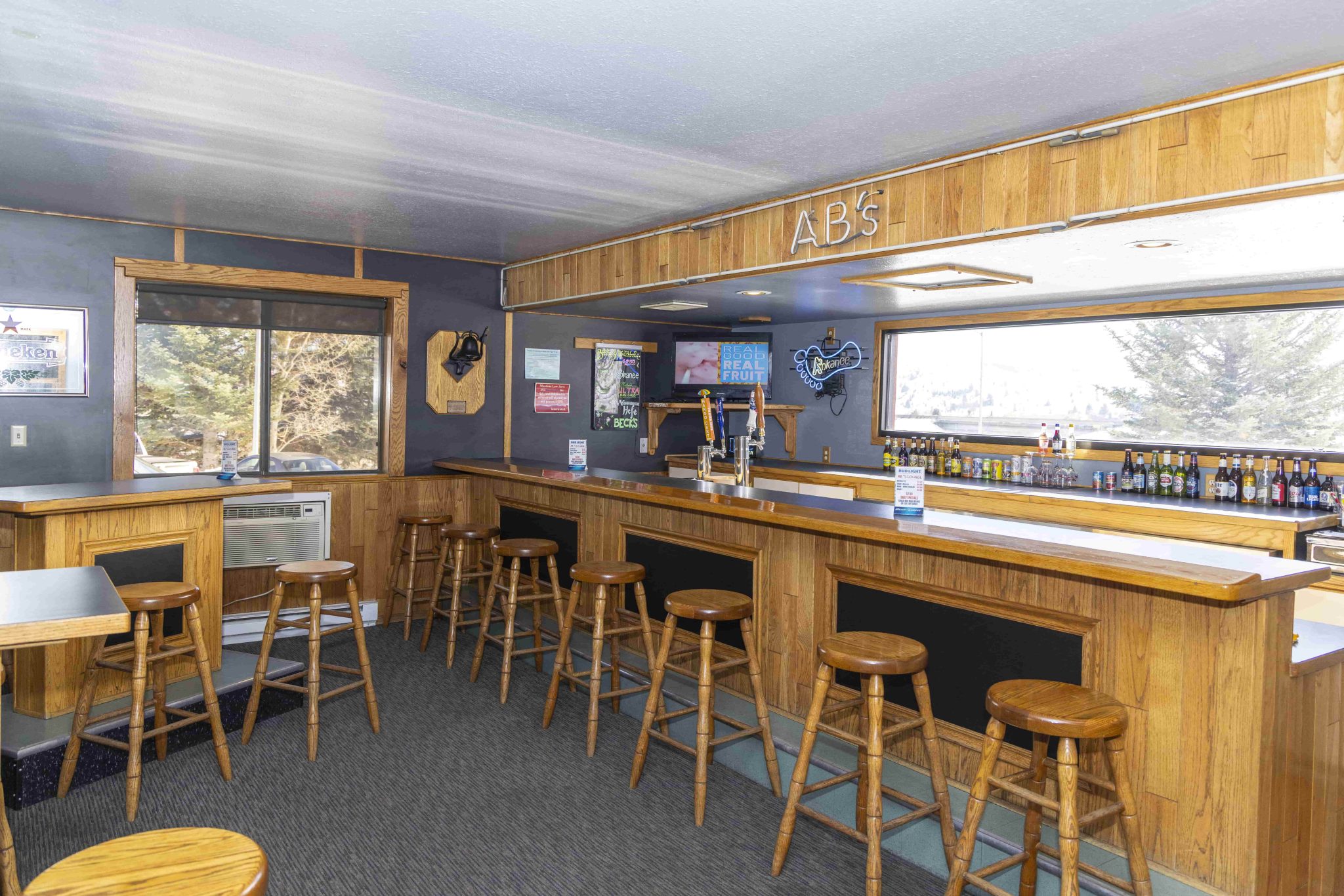 A bar with wooden counter and stools, showing a selection of beer bottles and a TV.