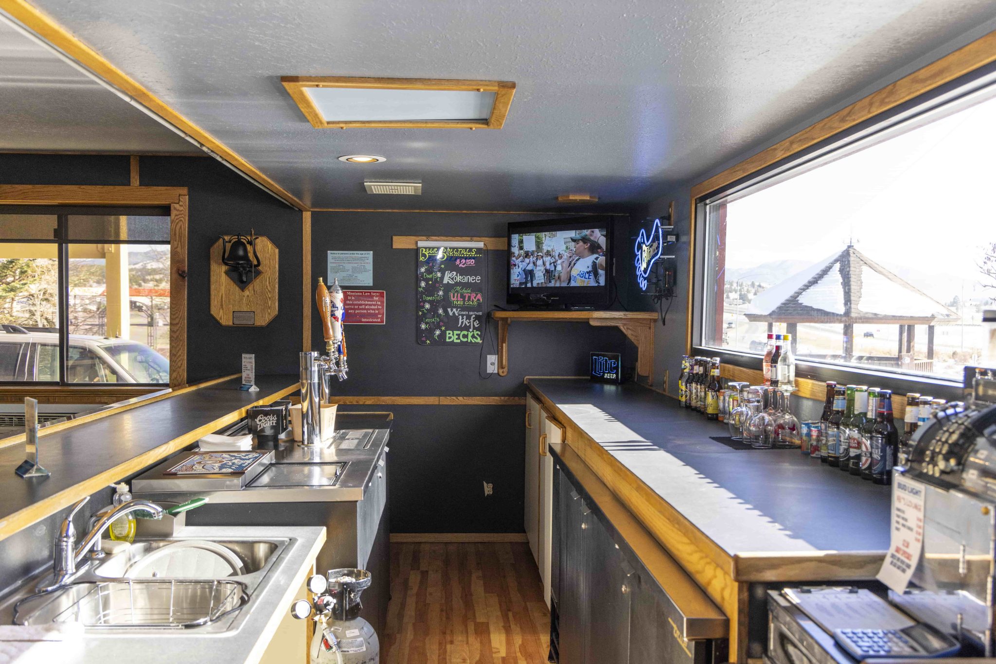 a bar with a tv, liquor, glasses, sink, and window