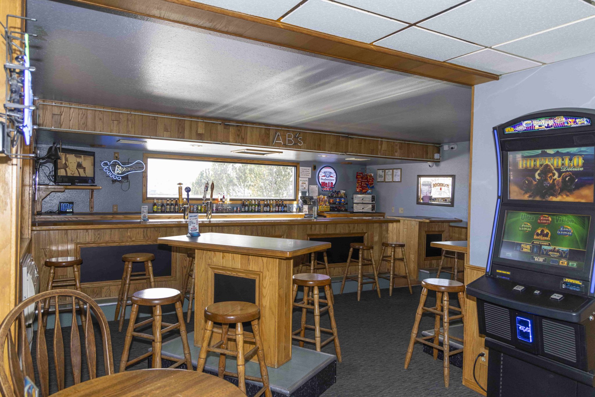 A dimly lit bar interior with a wooden bar, several stools, a gaming machine, and tables with chairs.